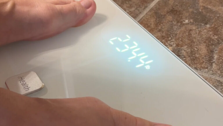 “iHealth Nexus smart scale on bathroom tile next to an iPhone showing weight trend