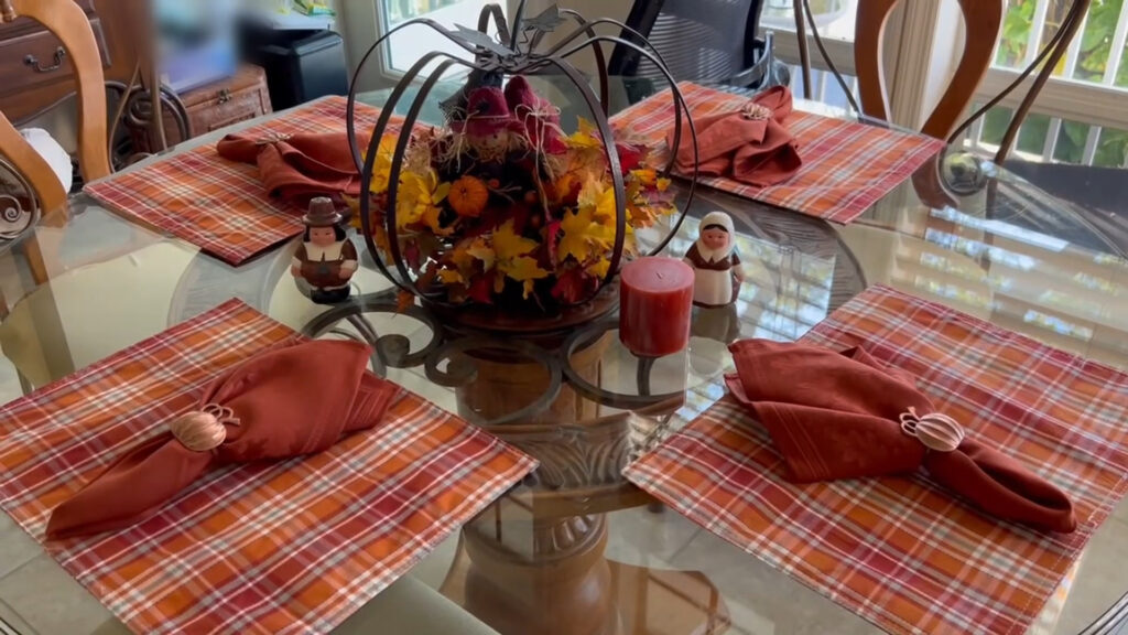 Fall dining table with buffalo plaid placemats, rusty orange cloth napkins in copper pumpkin napkin rings, Pilgrim figurines and fall centerpiece.